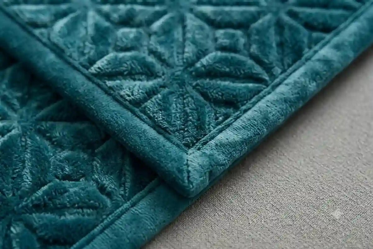 A close-up of a teal green quilted velvet mat with a geometric pattern and thick border, shown folded over on a neutral gray textured surface.