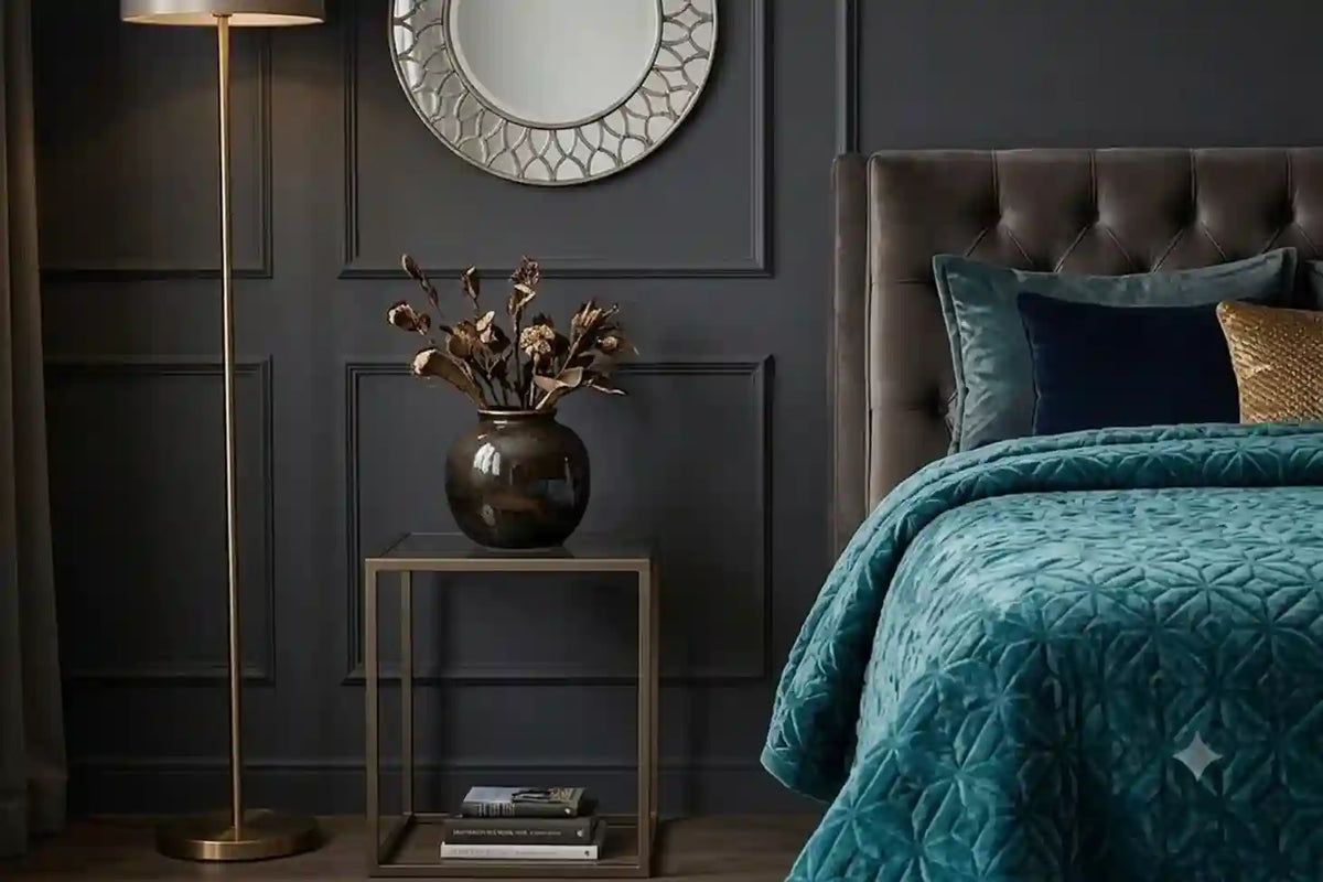 A styled bedroom corner featuring a bed with a textured teal quilt and dark pillows, next to a small side table with a vase of dried branches and a floor lamp against a dark paneled wall.