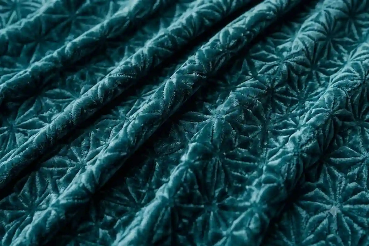 A close-up of a dark teal velvet fabric with a raised starburst or floral quilted texture, shown in soft folds.