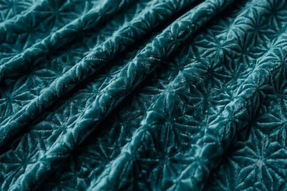 A close-up of a dark teal velvet fabric with a raised starburst or floral quilted texture, shown in soft folds.