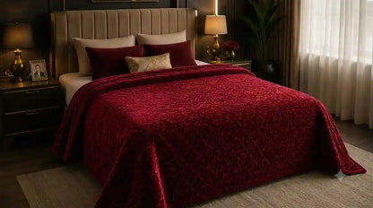 A bed dressed in a rich burgundy red textured quilted comforter with matching burgundy pillows, styled in a modern bedroom setting with a beige upholstered headboard, bedside tables, and warm ambient lighting.