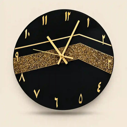 A round black wall clock with gold Arabic numeral hour markers, gold hands, and a wide glittery gold chevron stripe across the center, shown against a light beige background.