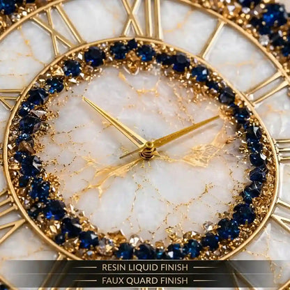 Close-up of a round wall clock with a gold-tone frame, Roman numerals, a white marbled dial, and a ring of deep blue faceted stones around the inner edge. Text at the bottom reads 'RESIN LIQUID FINISH' and 'FAUX QUARD FINISH'.
