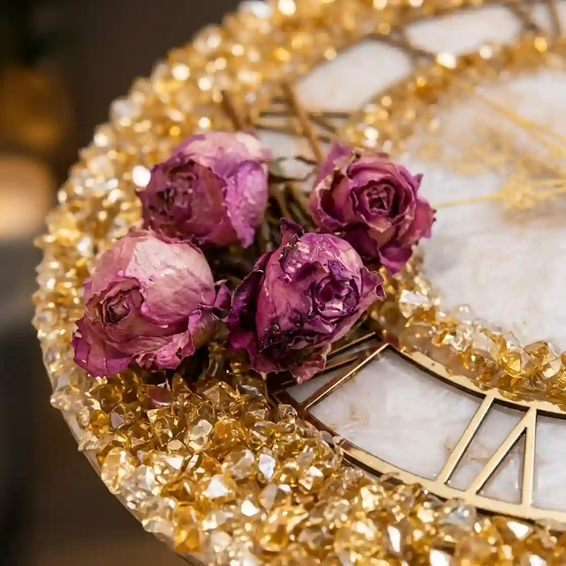 Four dried purple roses arranged on a round decorative tray with gold Roman numerals and a white marble inlay, surrounded by sparkling gold crystals.