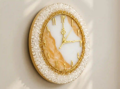 An oval wall clock with a gold-tone frame, gold hands, and gold Roman numerals, featuring a white and warm beige marbled dial and a sparkling clear crystal-like outer border, mounted on a light beige wall.
