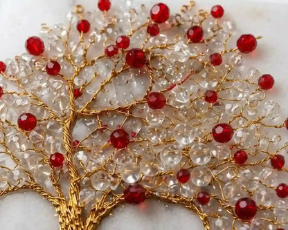 A close-up of a decorative tree design made of clear faceted beads on a gold wire frame, accented with multiple red faceted beads, set against a light neutral background.