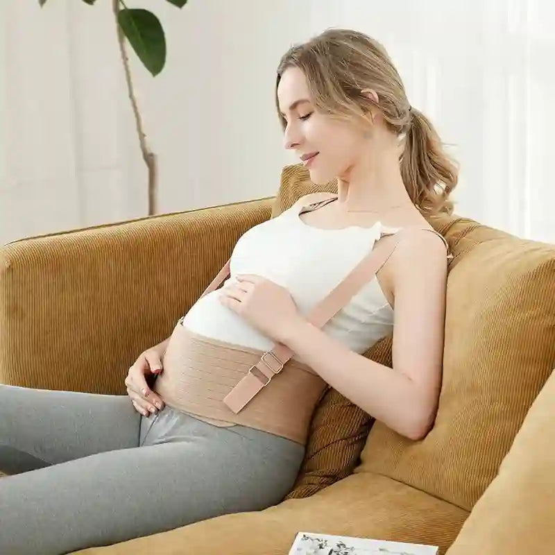 A pregnant woman wearing a beige maternity support belt sits on a brown corduroy couch in a bright, modern room.