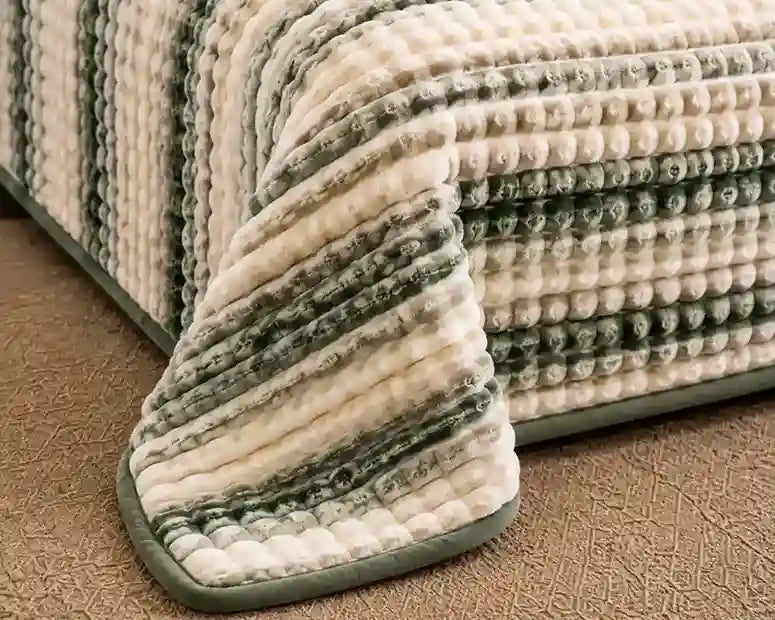 A close-up of a textured striped bath mat or rug draped over the corner of a bed, showing a plush ribbed pattern in cream, sage green, and gray on a brown carpeted floor.
