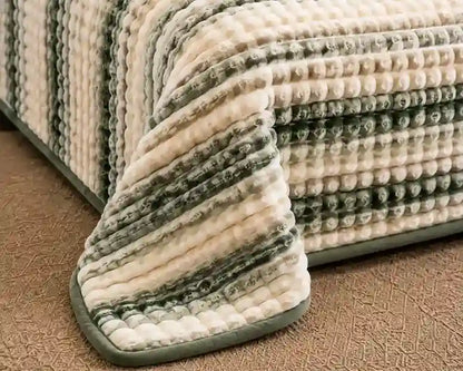 A close-up of a textured striped bath mat or rug draped over the corner of a bed, showing a plush ribbed pattern in cream, sage green, and gray on a brown carpeted floor.