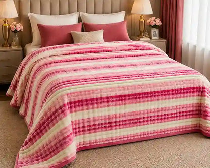 A bed dressed in a plush striped quilted blanket with horizontal bands of pink, magenta, and cream, styled with matching pink and beige pillows in a bedroom setting.