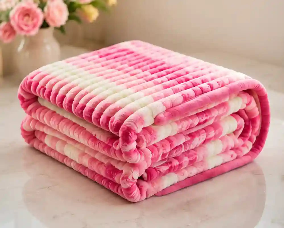 A folded plush blanket with a pink and white striped pattern, displayed on a light marble surface with a blurred vase of pink roses in the background.