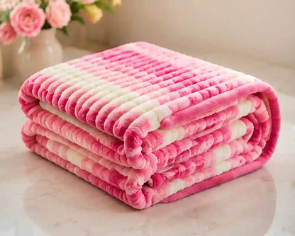 A folded plush blanket with a pink and white striped pattern, displayed on a light marble surface with a blurred vase of pink roses in the background.