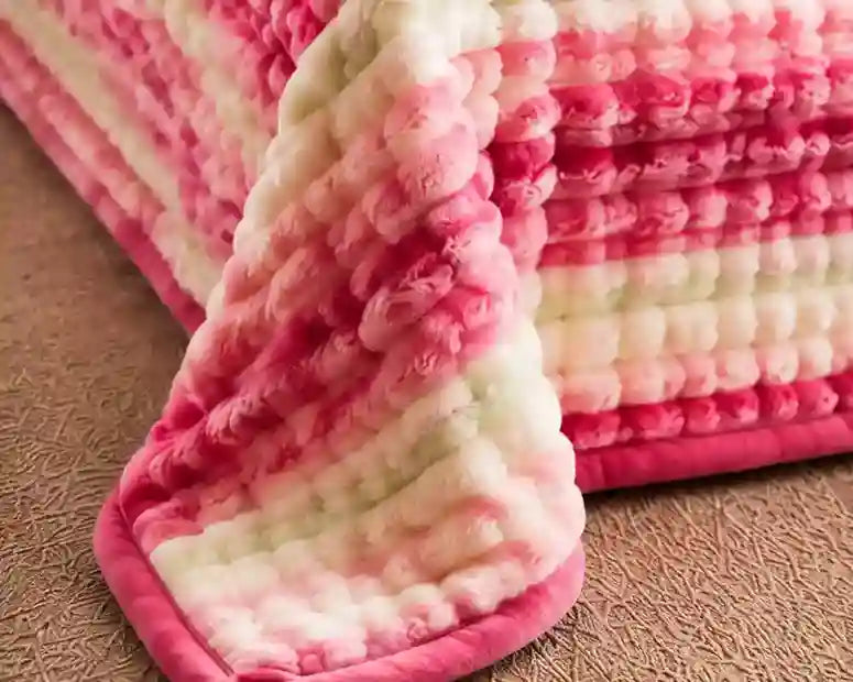 A close-up of a plush, chunky knit blanket with horizontal stripes in pink, magenta, and off-white, draped over the corner of a bed on a textured brown surface.