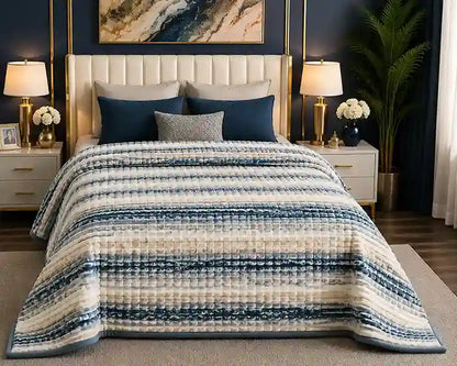 A bed dressed in a textured striped quilt in navy blue, cream, and gray, styled in a modern bedroom with matching pillows, bedside tables, lamps, and a dark blue wall.