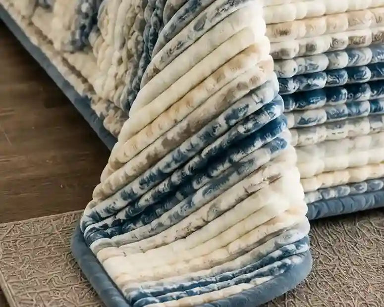 A close-up of a plush, ribbed bath mat or rug corner in cream and slate blue, shown folded to display its soft texture and thick padding, with a matching stack of mats in the background.