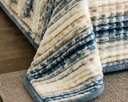 A close-up of a plush, ribbed bath mat or rug corner in cream and slate blue, shown folded to display its soft texture and thick padding, with a matching stack of mats in the background.