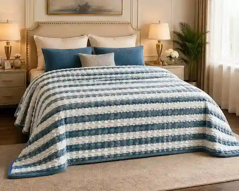 A striped quilted blanket in shades of blue and white is styled on a bed in a warm, elegant bedroom setting with matching pillows, bedside tables, lamps, and a plant.