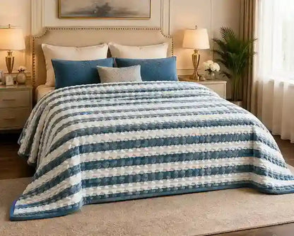 A striped quilted blanket in shades of blue and white is styled on a bed in a warm, elegant bedroom setting with matching pillows, bedside tables, lamps, and a plant.