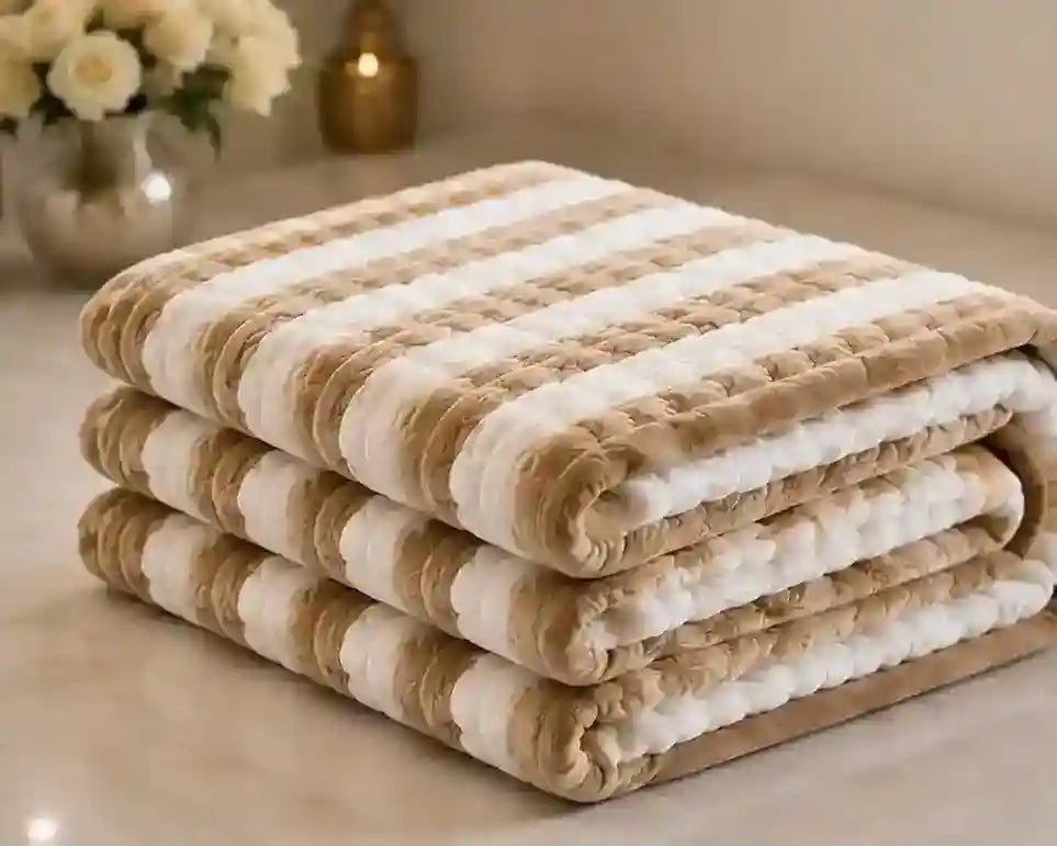 Three folded plush striped blankets in beige and white, stacked on a light beige surface with a softly blurred background showing flowers and a candle.
