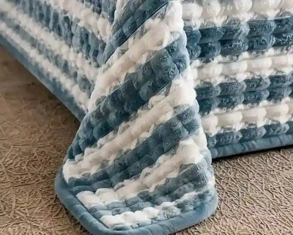 A close-up of a textured, chunky striped blanket or quilt in blue and white, draped over the corner of a bed on a beige carpeted floor.