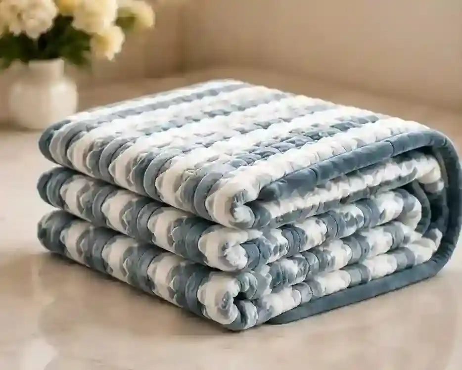 A folded plush blanket with a blue and white cloud-like textured pattern, displayed on a light beige surface with a soft-focus background.
