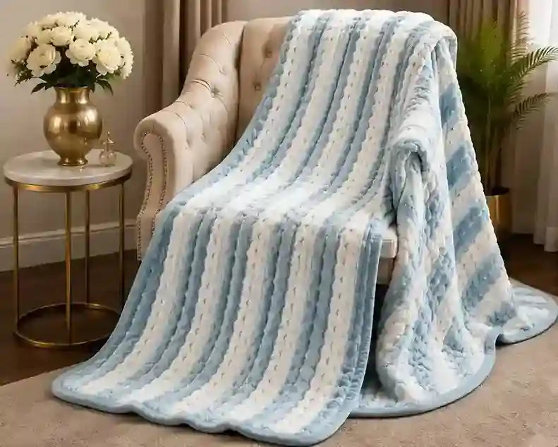 A plush striped throw blanket in light blue and white is draped over a cream tufted armchair in a styled living room setting with a side table and flowers.
