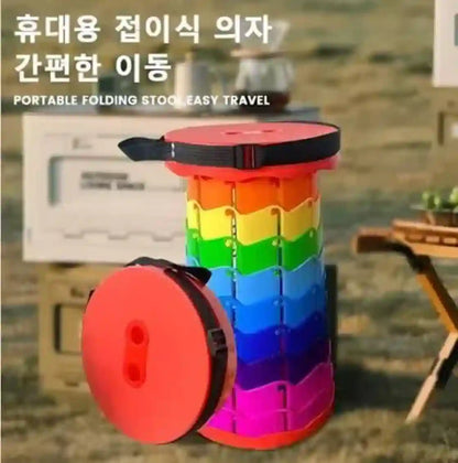 A portable folding camp stool with a rainbow-colored striped body and red circular seat/top, shown outdoors with Korean text on the image. The stool is open and photographed in a blurred campsite-like environment.