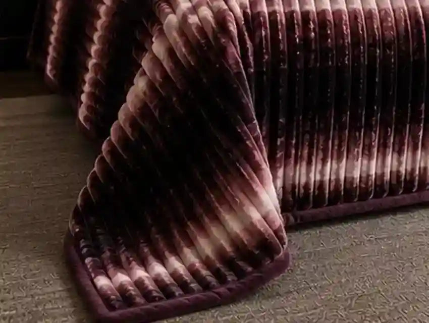A close-up of a plush, ribbed blanket or throw draped over the corner of a bed, featuring a deep plum purple and dark brown tie-dye pattern with lighter cream highlights.