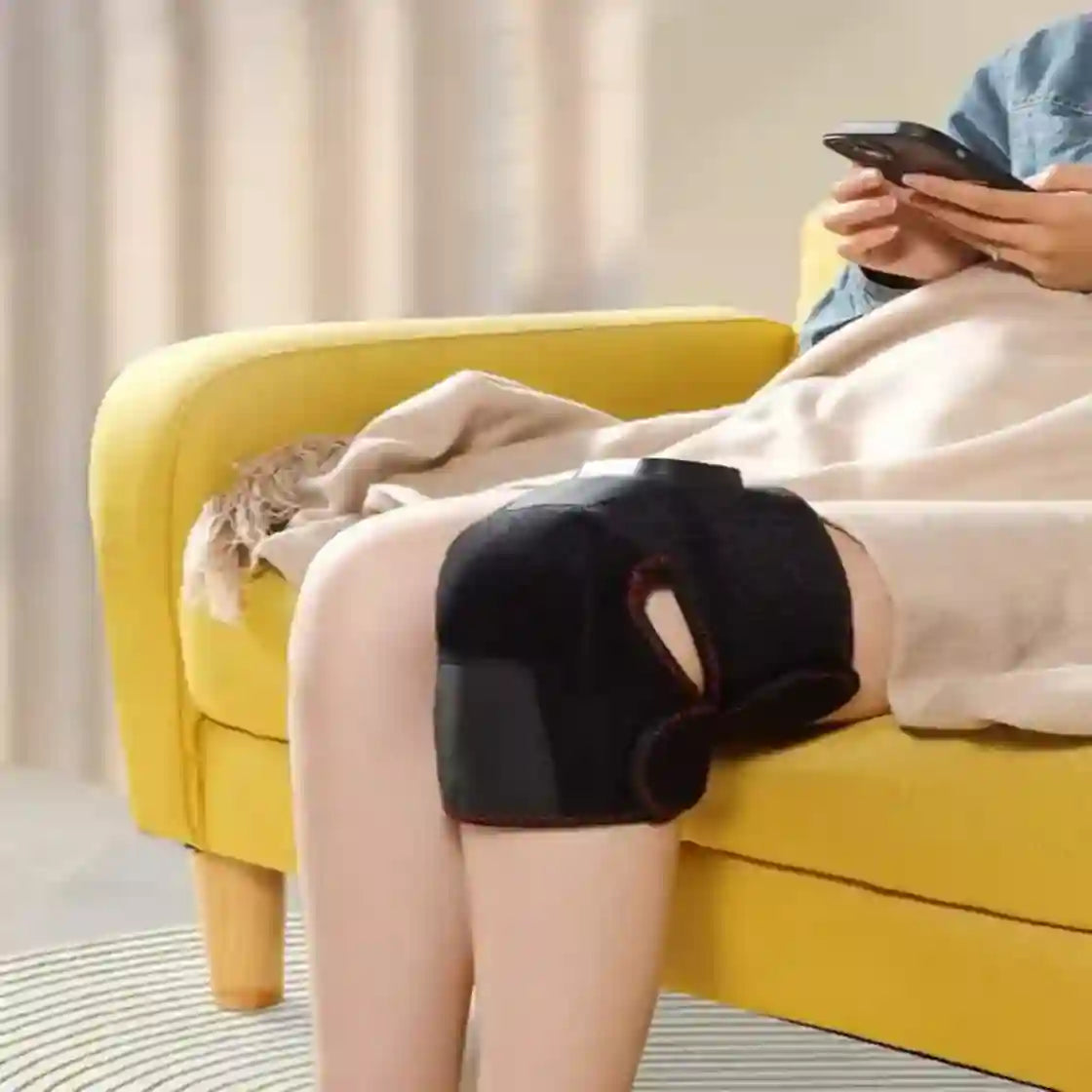 Person sitting on a yellow sofa using a smartphone with a blanket and pillow nearby.