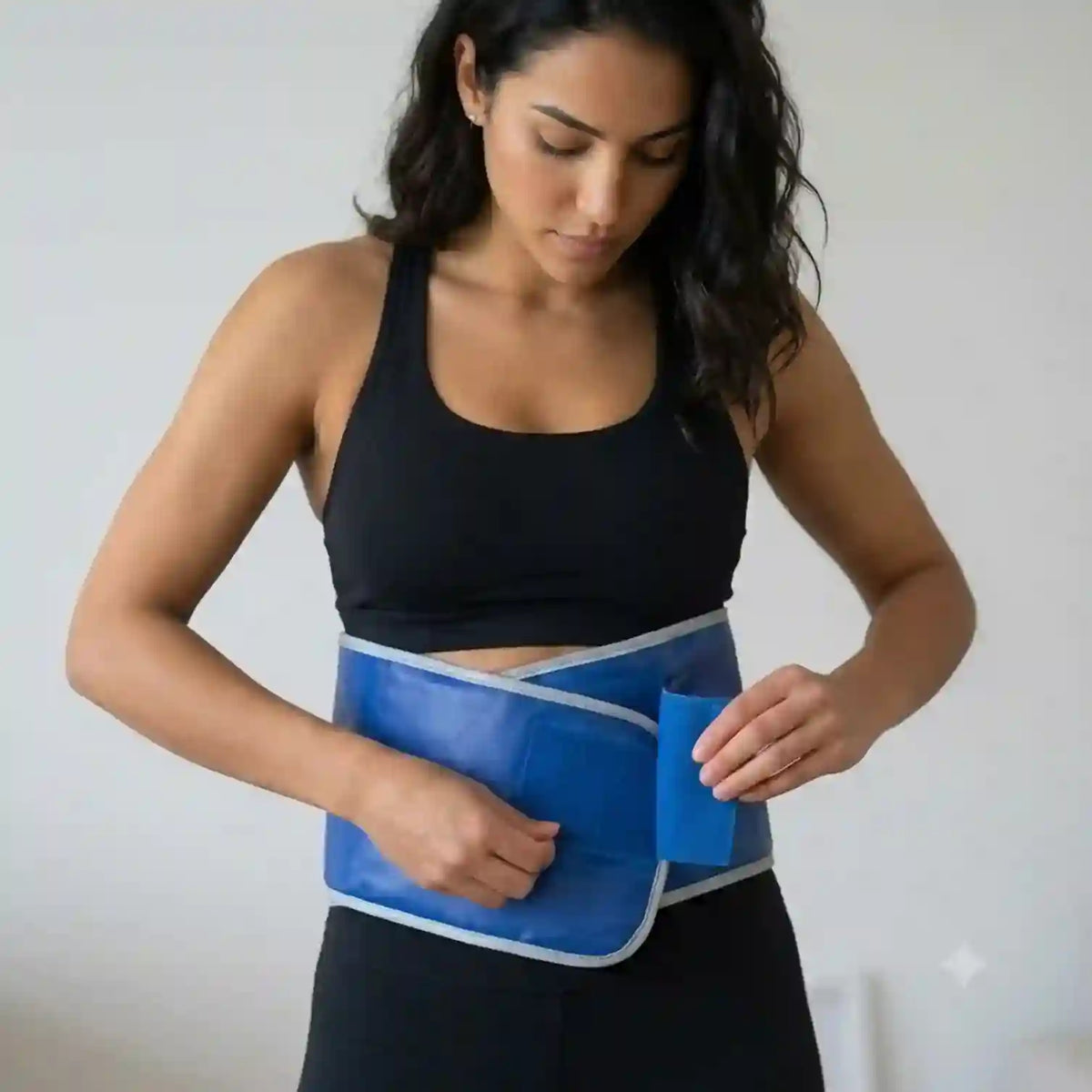 A woman wearing a black sports bra and black leggings is shown from the waist up, wrapping a blue abdominal support belt with white trim around her midsection.