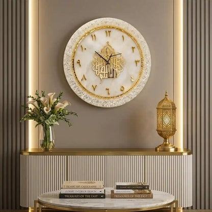 A round Islamic wall clock with gold Arabic calligraphy and gold Arabic numerals on a white marble-patterned face, mounted on a gray wall above a console table decorated with a vase of flowers and a gold lantern.