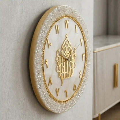 An oval wall clock with a gold frame, white marble-look face, Arabic numeral hour markers, and a decorative gold central motif, mounted on a light gray wall next to a white cabinet.