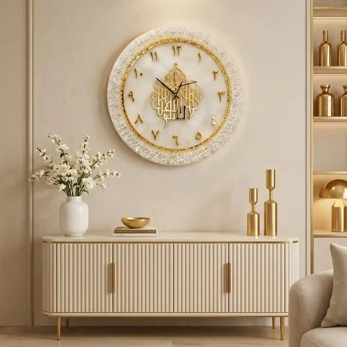 An ornate round wall clock with a white textured frame and gold Arabic numerals, mounted on a beige wall above a cream-colored ribbed console table decorated with a vase of white flowers and gold candle holders.