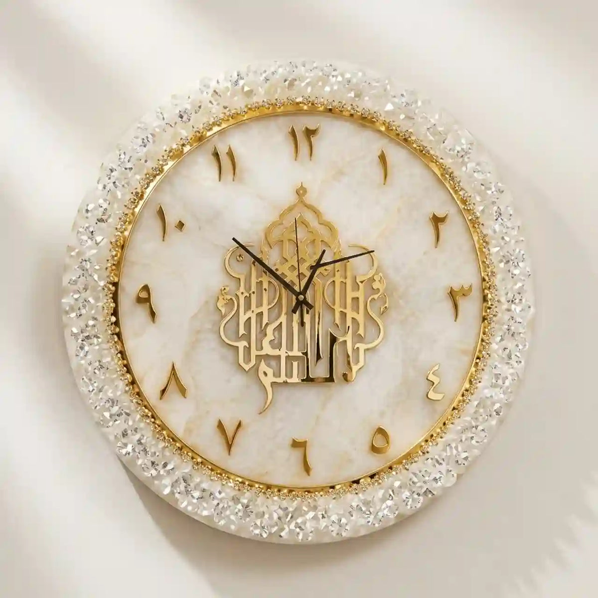 An ornate round wall clock with a white marble face, a gold Arabic calligraphy center design, gold Arabic numerals, and a crystal-embellished border, shown against a light neutral background.