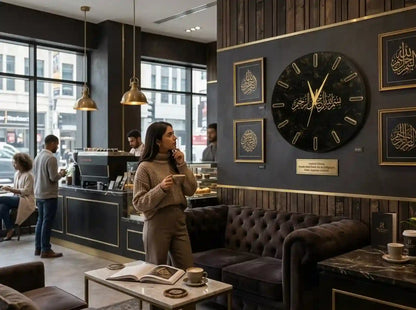 A large black wall clock with gold Arabic calligraphy hands and markers is mounted on a dark gray wall in a modern cafe setting. The scene includes a woman standing nearby, a tufted brown sofa, a coffee counter, and framed gold decorative art.