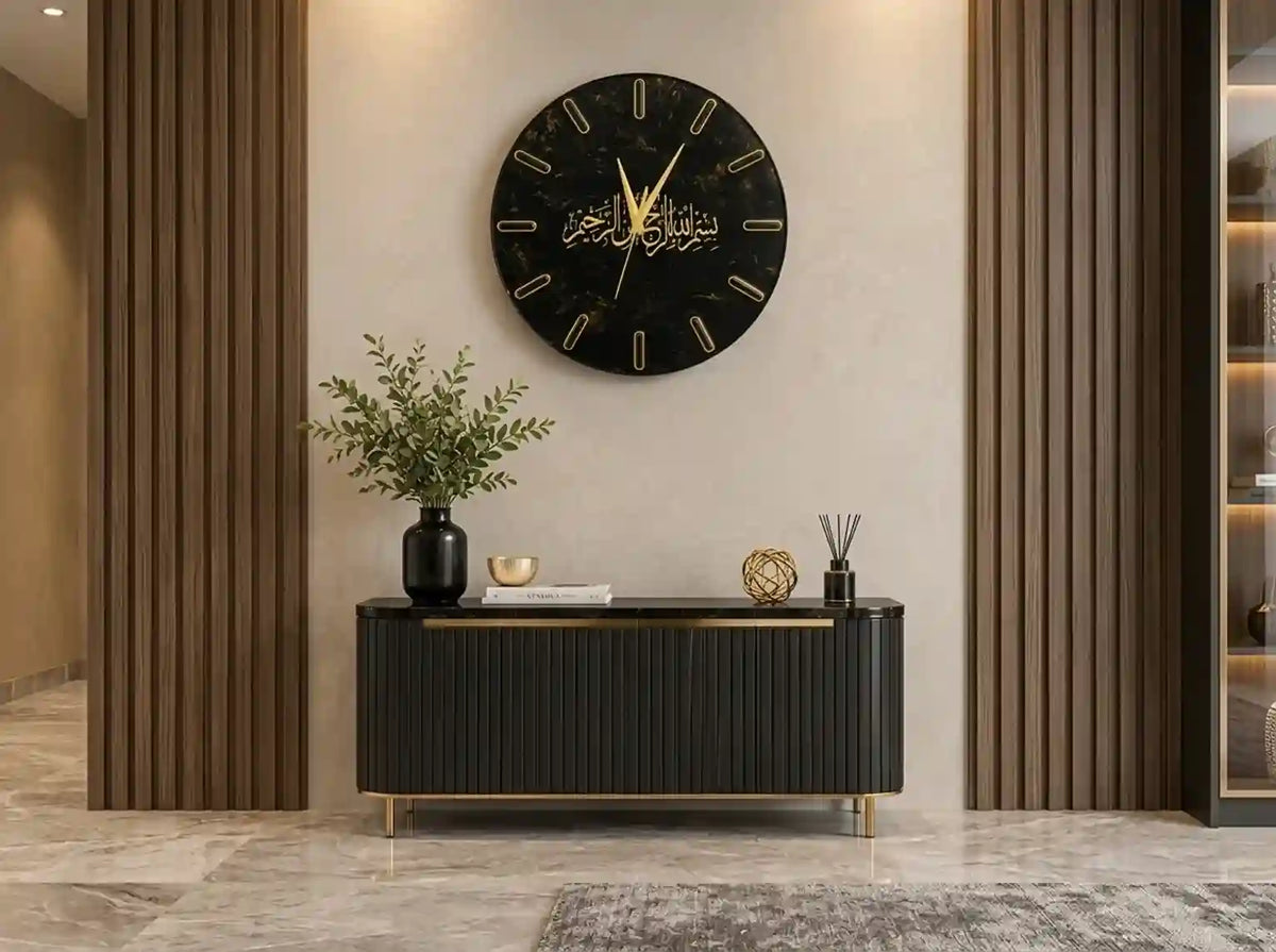 A round black wall clock with gold Arabic numerals and hands, mounted on a beige wall above a black console table with gold accents in a modern interior setting.