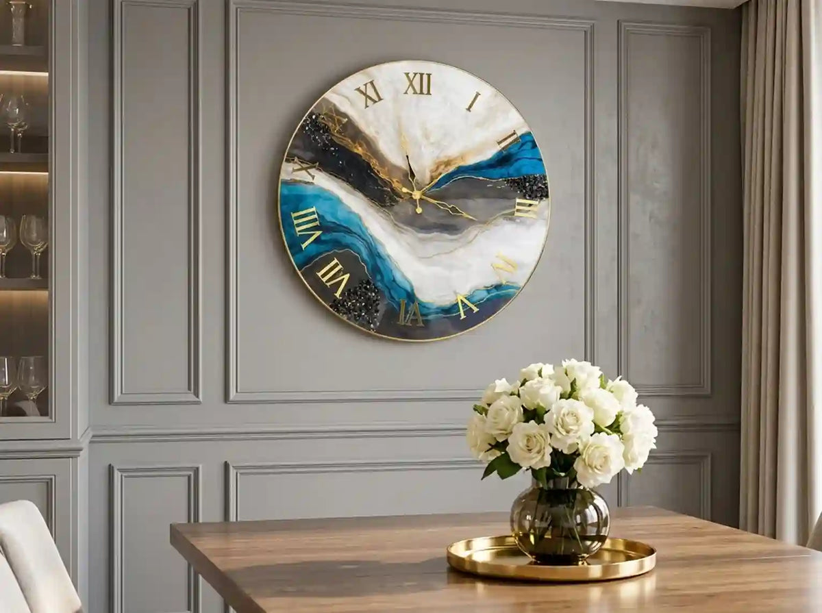 Round wall clock with an abstract blue, white, and black marbled design and gold Roman numerals, mounted on a gray paneled wall above a wooden table with white flowers.