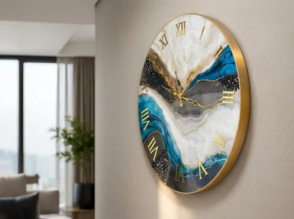 A round wall clock with a gold frame and gold Roman numerals, featuring an abstract marbled pattern in white, blue, black, and gray, mounted on a beige wall in a modern living room setting.