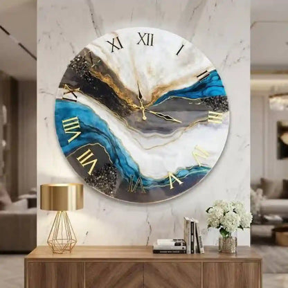 A large round wall clock with an abstract marbled design in blue, white, black, and gold, featuring gold Roman numerals. The clock is mounted on a white marble-patterned wall above a wooden console table with a gold lamp and decorative items.