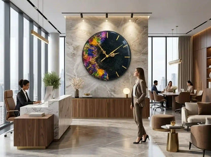 A large round wall clock with a colorful abstract watercolor design on a black background, mounted on a marble-patterned wall in a modern office lobby. Businesspeople are seen working and standing in the lobby around the clock.