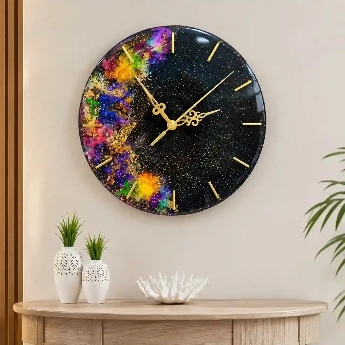 A round wall clock with a black glittery space-like background and a colorful abstract floral burst on the left side, mounted on a beige wall above a light wood console table with decorative vases and a plant.