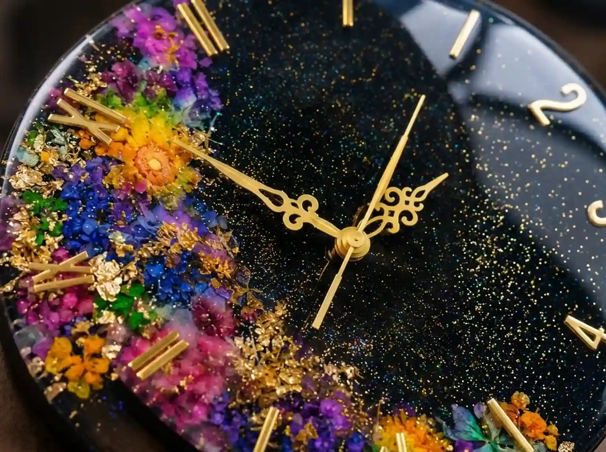 A close-up of a decorative round clock face with a black glittery background, gold hands, gold Arabic numeral hour markers, and embedded multicolored dried flowers and gold leaf accents.