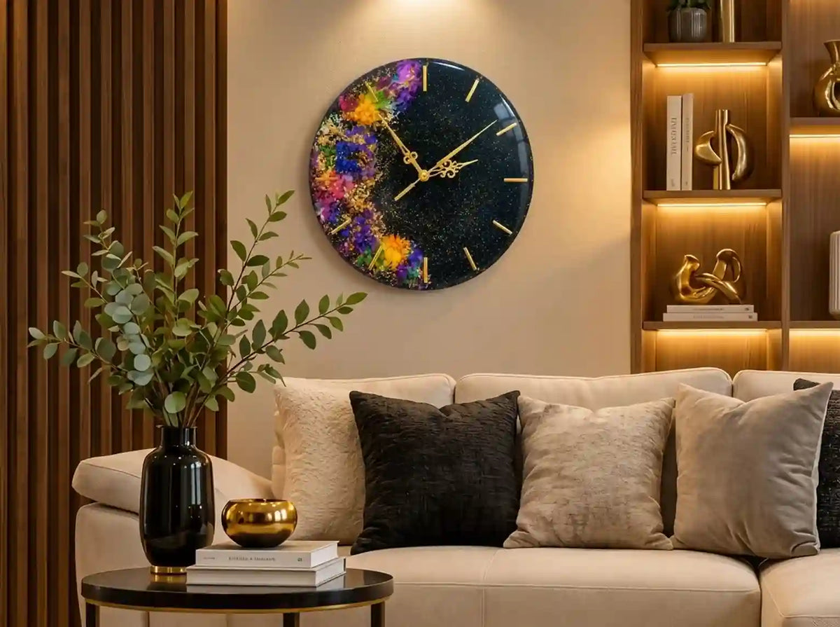 A round wall clock with a colorful cosmic paint-splatter design on a black background, featuring gold-tone hands, mounted on a beige wall above a modern sofa in a styled living room.