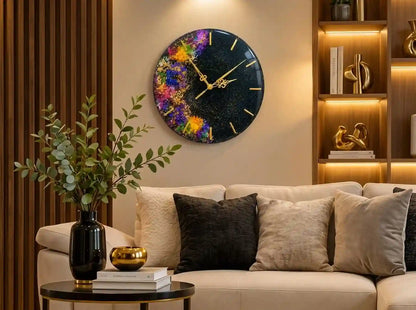A round wall clock with a colorful cosmic paint-splatter design on a black background, featuring gold-tone hands, mounted on a beige wall above a modern sofa in a styled living room.