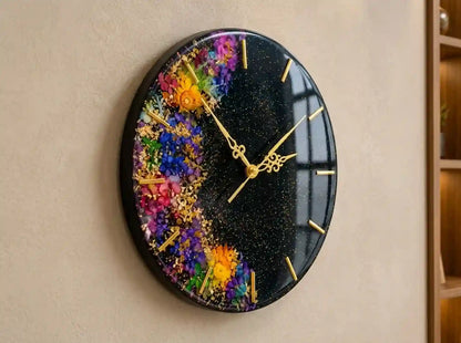 An oval wall clock with a glossy black background, embedded with colorful dried flowers and gold glitter. The clock features gold hands and gold hour markers, mounted on a beige textured wall.