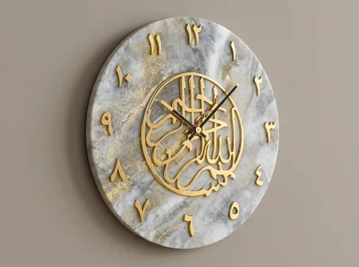 Round wall clock with a white and gray marble-look face, gold Arabic numeral symbols, and a central gold circular calligraphic design, shown against a plain taupe wall.