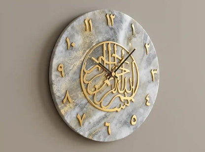 Round wall clock with a white and gray marble-look face, gold Arabic numeral symbols, and a central gold circular calligraphic design, shown against a plain taupe wall.
