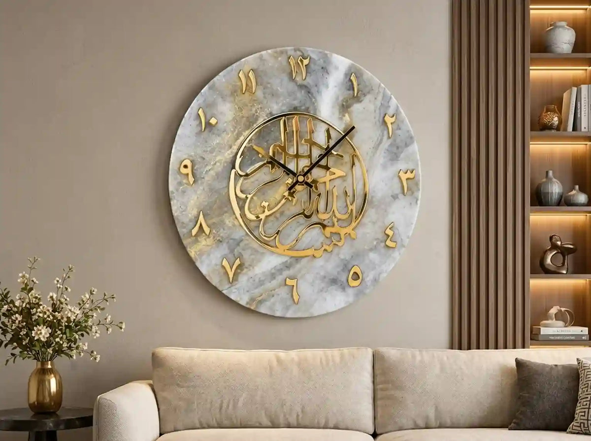 A large round wall clock with a gray and white marble pattern face, gold Arabic numerals, and a gold central calligraphic design, mounted on a beige wall above a light beige sofa in a modern living room setting.