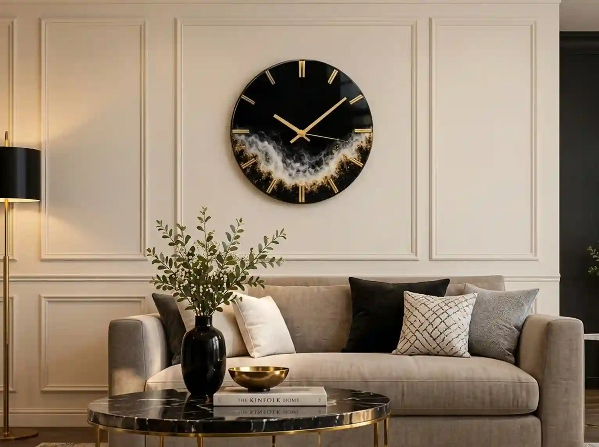 A round black wall clock with gold Roman numerals and gold hands, featuring a gold and white smoky wave design at the bottom, mounted on a cream paneled wall above a beige sofa.