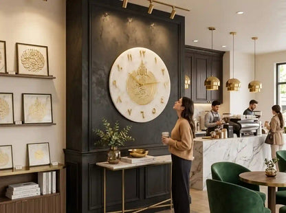 A large circular white and gold wall clock with Roman numerals is mounted on a dark paneled wall in a modern cafe interior. A woman stands in front of the clock holding a cup, while coffee bar staff work behind a marble counter with green chairs in the foreground.